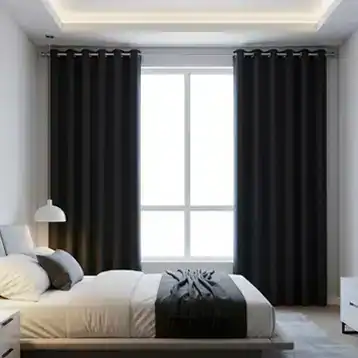A modern bedroom with a neatly made bed, white bedding, and a black throw blanket. There is a large window with black curtains, a bedside table with a lamp, and minimalistic white walls and decor.