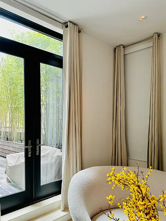 A bright room features tall white curtains, black-framed glass doors leading to an outdoor patio with green bamboo, a curved white chair, and a vase of yellow flowers on a table in the foreground.