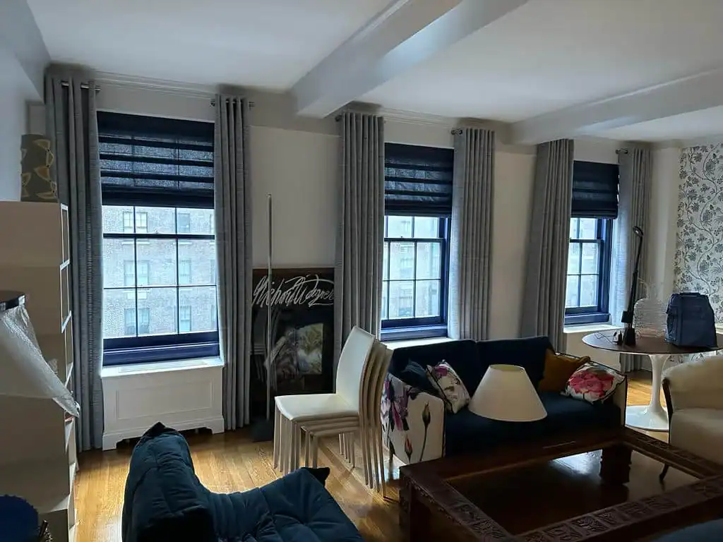 A living room with wooden floors, a dark blue sofa, stacked white chairs, a coffee table with a lamp, and large windows with gray curtains and dark blinds, letting in natural light.