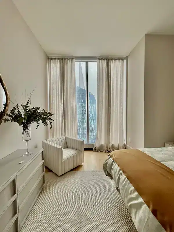 A bright, modern bedroom with a large window, sheer curtains, a white armchair, a dresser with a vase of greenery, and a bed with beige and white bedding. A cityscape is visible outside the window.