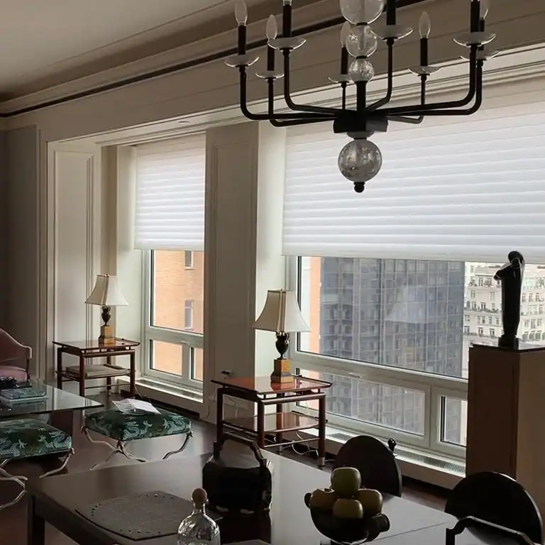 A bright, modern living room with large windows covered by white blinds, elegant furniture, table lamps, a black chandelier, and a cityscape visible outside. A table with fruit and glassware is in the foreground.