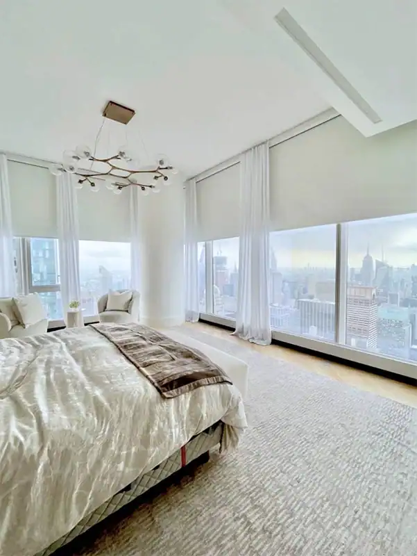 Spacious, modern bedroom with large windows offering a panoramic city skyline view. The room features a neatly made bed, two armchairs, light-colored curtains, and a contemporary chandelier.