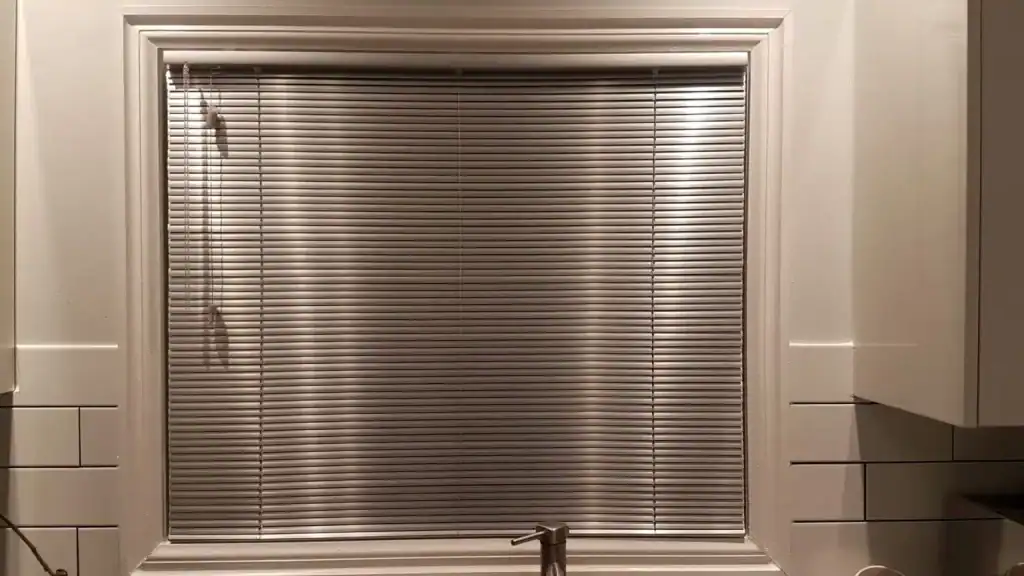 A closed silver horizontal window blind with a pull cord, framed by white trim. A faucet is visible at the bottom center, indicating the window is above a sink.