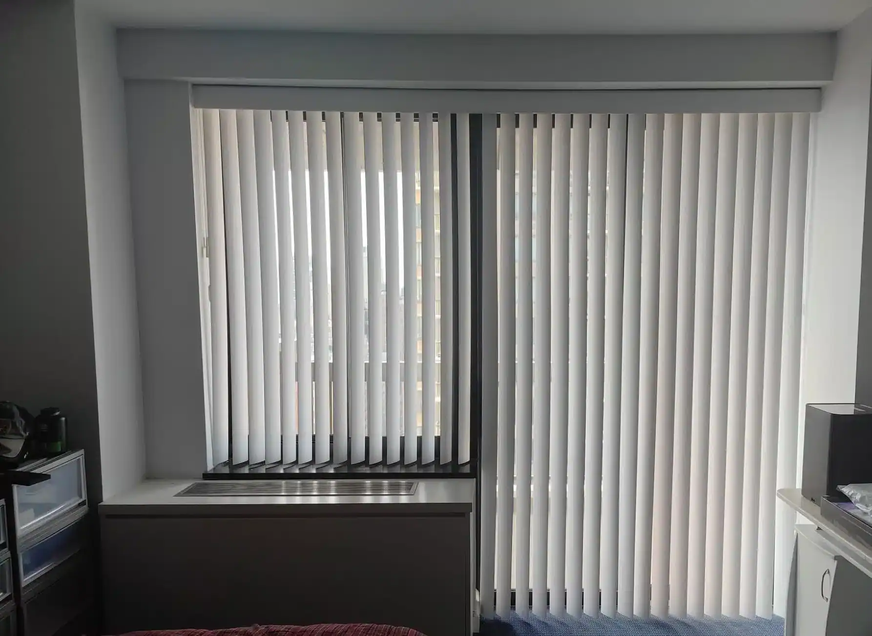 A large window with vertical white blinds, partially open to let in daylight, brightens this FL home. Below the window is a radiator cover, while parts of a bed and other furniture showcase thoughtful design by a top home design company in Miami-Dade & Broward County.