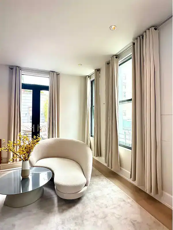 A modern, bright living room with large windows covered by beige curtains, a curved white sofa, a round glass coffee table, and a vase with yellow flowers on the table.