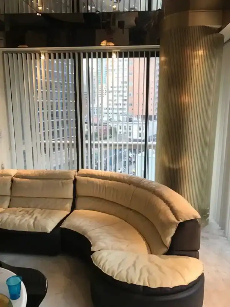 A modern living room with a curved beige sectional sofa, floor-to-ceiling windows with vertical blinds, city buildings visible outside, and a tall ribbed pillar next to the windows.