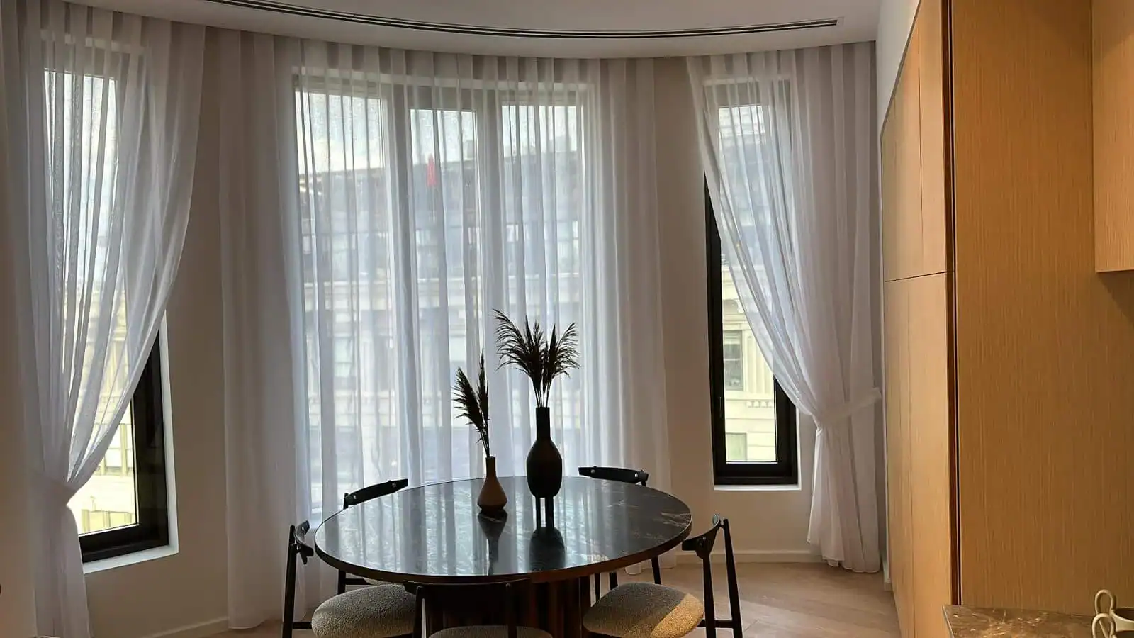 A modern dining area with a round black table, four chairs, and decorative vases, set by large windows covered with sheer white curtains that let in natural light.