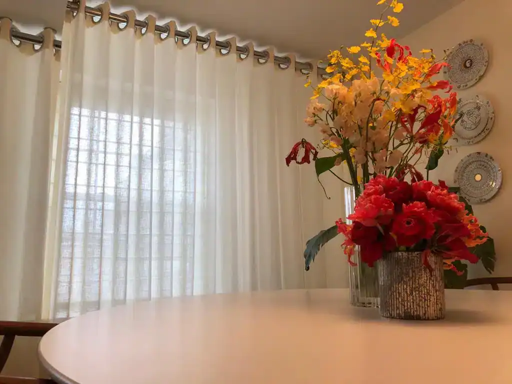 A round table with red and yellow flower arrangements in vases sits near a window with sheer white curtains. Decorative plates hang on the beige wall in the background.