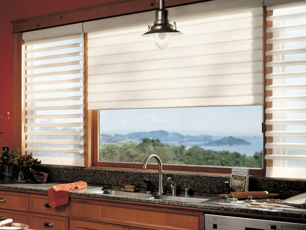 A kitchen with a granite countertop, sink, and wooden cabinets. Large windows with white blinds offer a view of green trees and distant blue mountains. A metallic pendant light hangs above the sink.