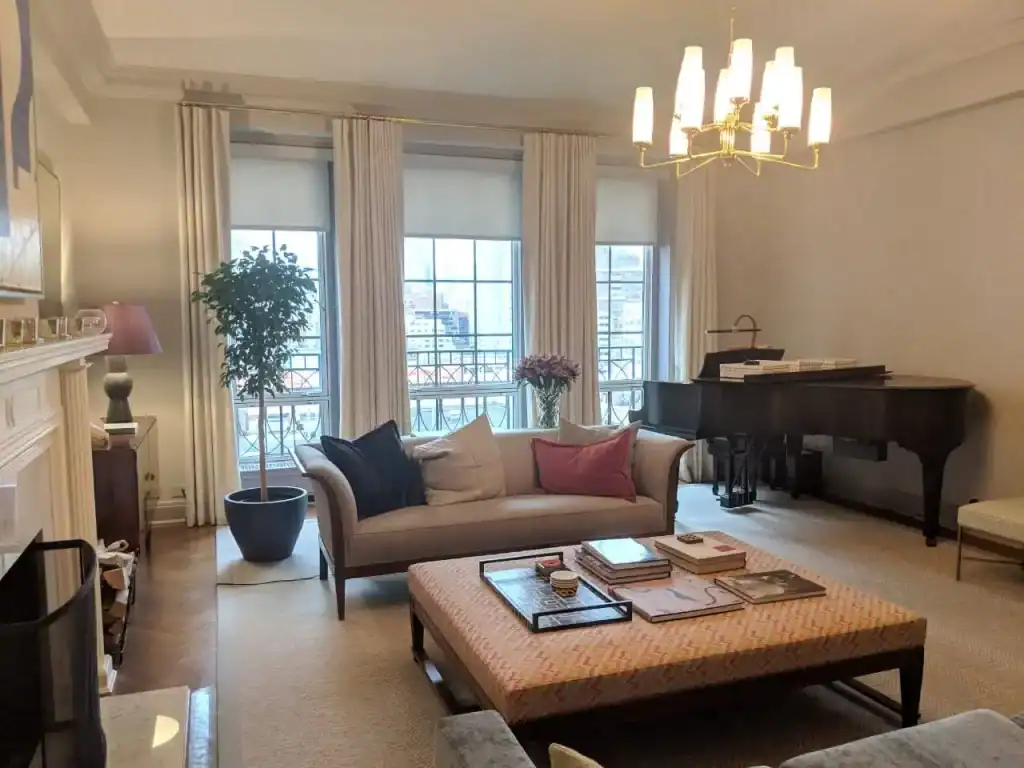 A bright living room with large windows, white curtains, a beige sofa with assorted pillows, a patterned ottoman, a grand piano, a green potted plant, and a modern chandelier hanging from the ceiling.