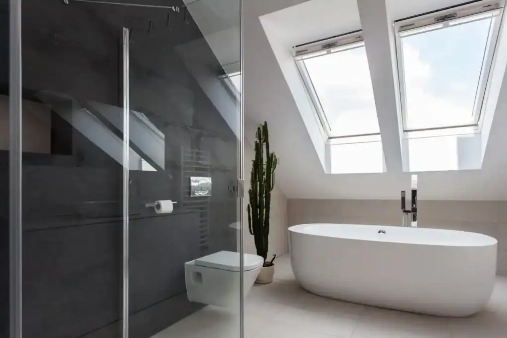 A modern bathroom with a freestanding white bathtub beneath two large skylights, a glass-enclosed shower, a wall-mounted toilet, dark accent walls, and a tall potted cactus.