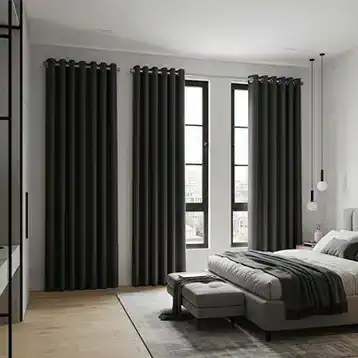 Modern bedroom with large windows covered by dark curtains, a neatly made bed with gray bedding, a small bench at the foot of the bed, pendant lights, and light wood flooring.