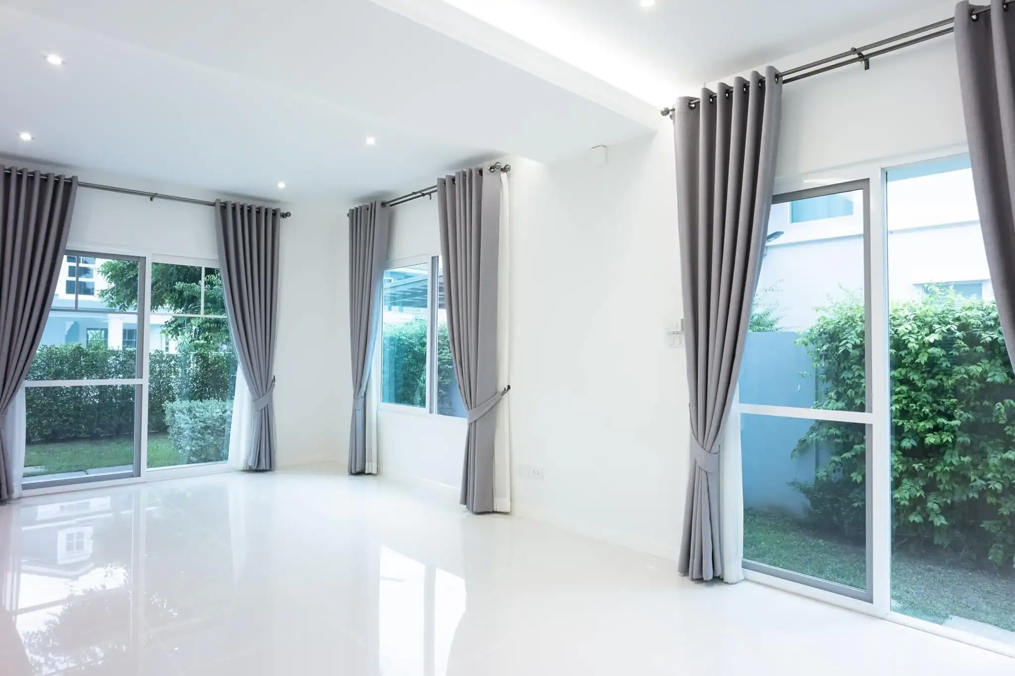 A bright, empty room with large windows and glass doors, framed by light gray curtains. The white walls and floor reflect natural light, with green bushes outside—a perfect space crafted by a top home design company in Miami-Dade & Broward County, FL.