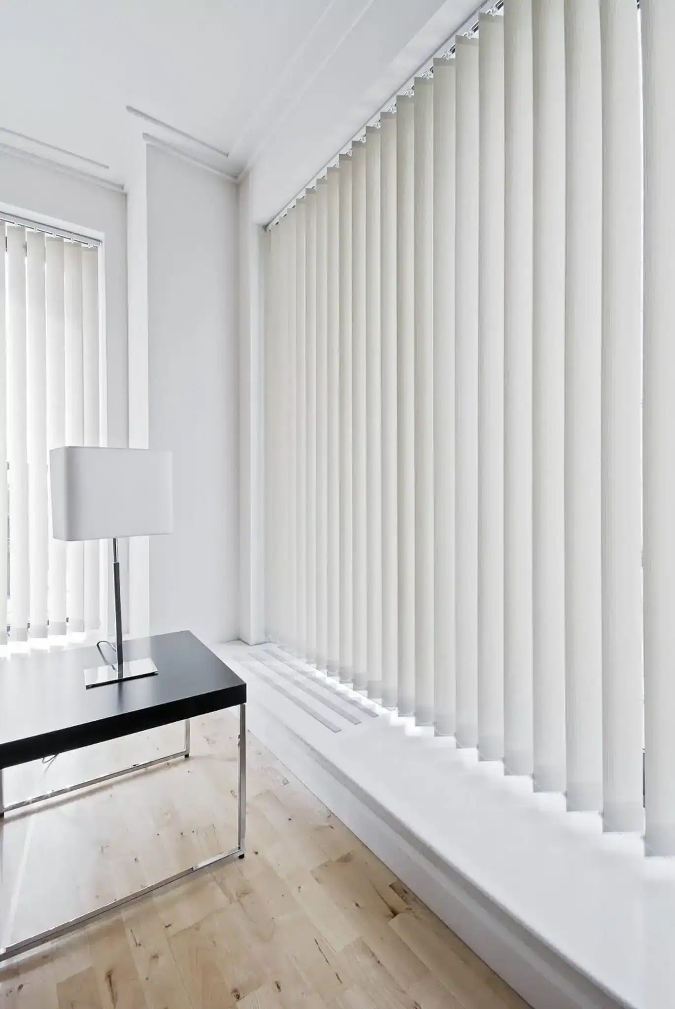 A modern, minimalistic office corner with a black desk, a white table lamp, and large windows covered by vertical white blinds. The space features light wood flooring and white walls.