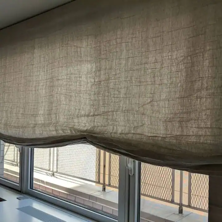 A beige linen curtain from a top home design company Miami-Dade & Broward County partially covers a window. The lower part is gathered and slightly raised, revealing a metal fence and part of the building exterior outside.