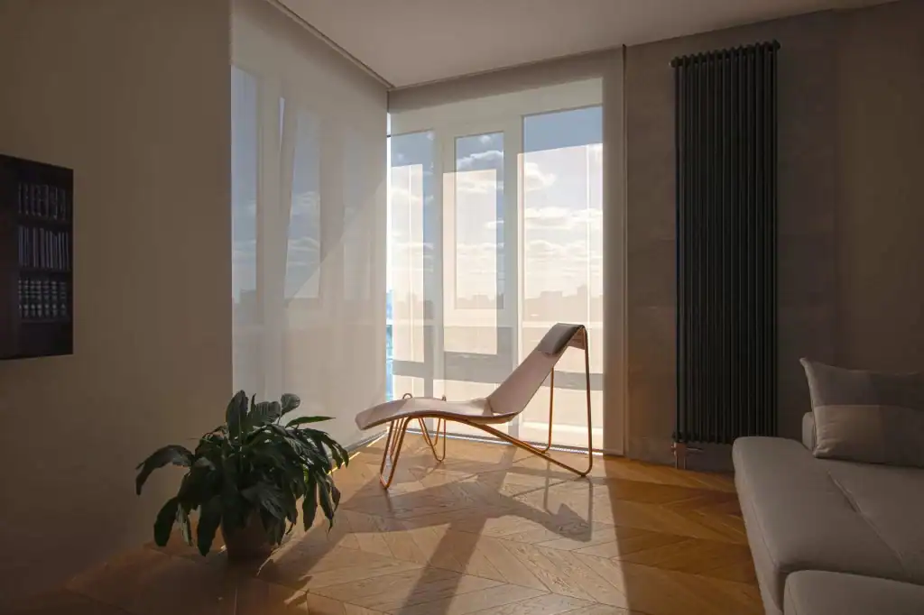 A sunlit modern living room with a lounge chair by floor-to-ceiling windows, sheer curtains, a tall black radiator, a green potted plant, and part of a gray sofa on a herringbone wooden floor.