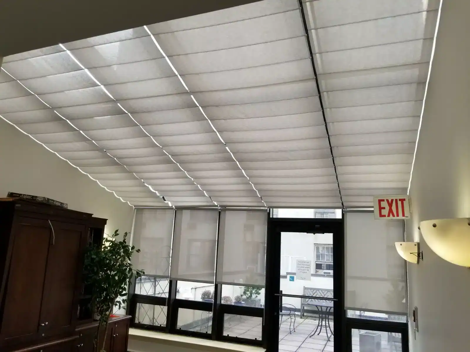 A slanted ceiling window with white fabric blinds partially covering the glass. Below are large windows and a glass door leading outside; a red exit sign and some indoor furniture are visible.