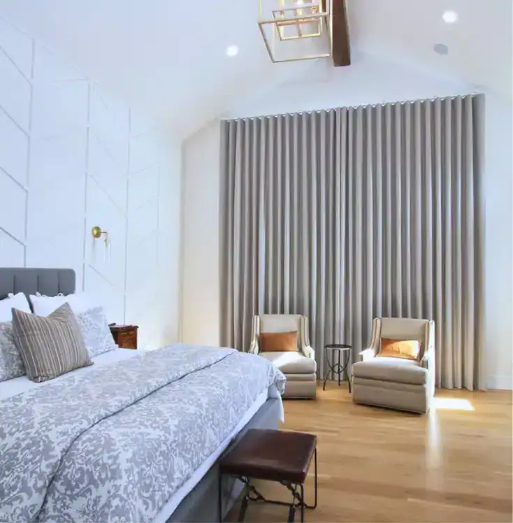 A modern bedroom with a large bed, patterned gray bedding, two beige armchairs with pillows, a small side table, floor-length gray curtains, and wooden floors under a vaulted ceiling with a wooden beam.