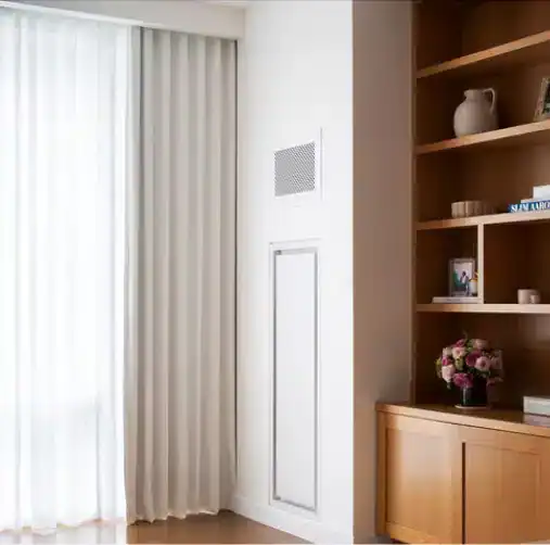 A bright, modern living space by a top home design company Miami-Dade & Broward County, with sheer white curtains, a built-in wooden bookshelf holding books, a vase, framed photo, and flowers beside a white vent panel.