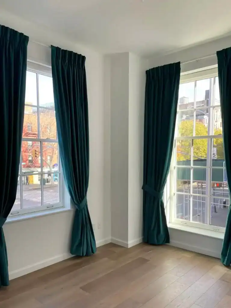 A bright corner room with two large windows, each framed by dark green curtains. Sunlight streams in, highlighting the light walls and medium-tone wooden floor. Trees and buildings are visible outside.