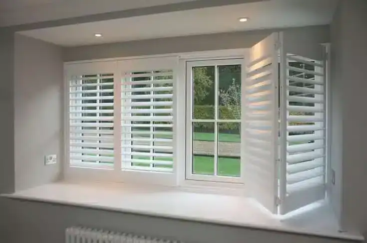 A white-framed window with plantation shutters; two shutters are closed while one on the right is open, revealing a green outdoor landscape. The window sits in a recessed wall with a wide windowsill.