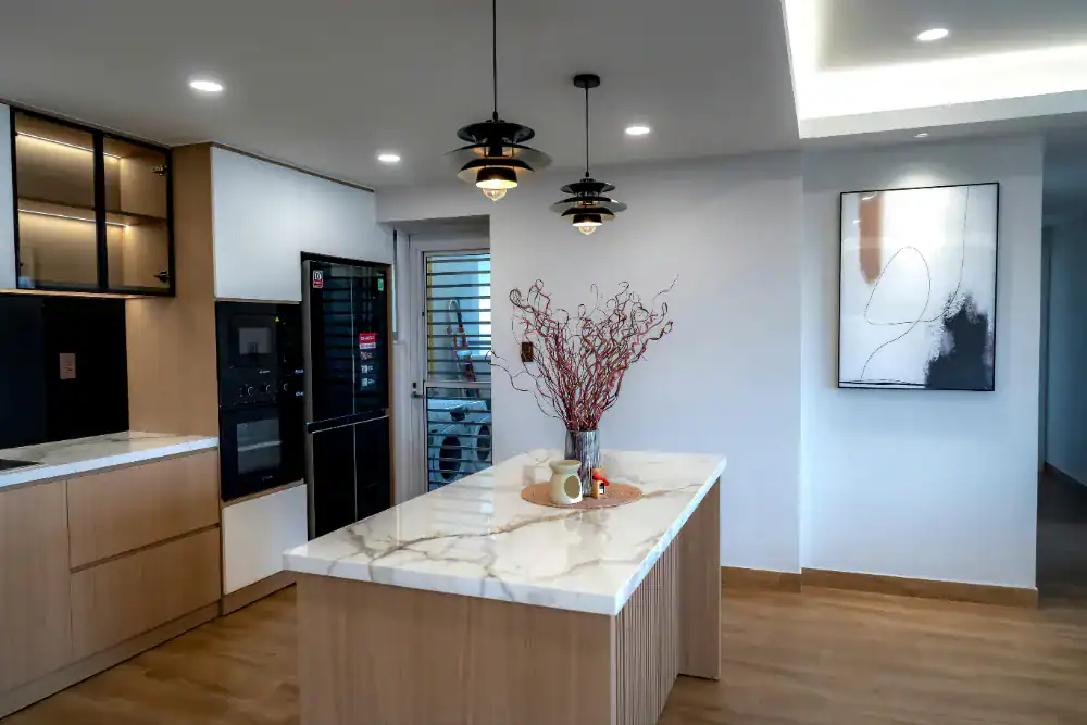 Modern kitchen with light wood cabinets, a marble island countertop, pendant lights, and a centerpiece vase with dried branches. There is minimal decor, a black refrigerator, and abstract art on a white wall.