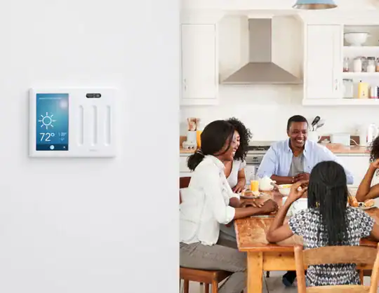 A smart thermostat displays 72°F and sunny on a wall in a bright kitchen, while four people sit together at a wooden table, smiling and talking over breakfast.