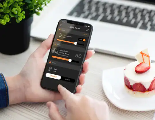 A person uses a smartphone app to control smart home devices, adjusting lights and temperature, with a dessert and plant on a white table in the background.