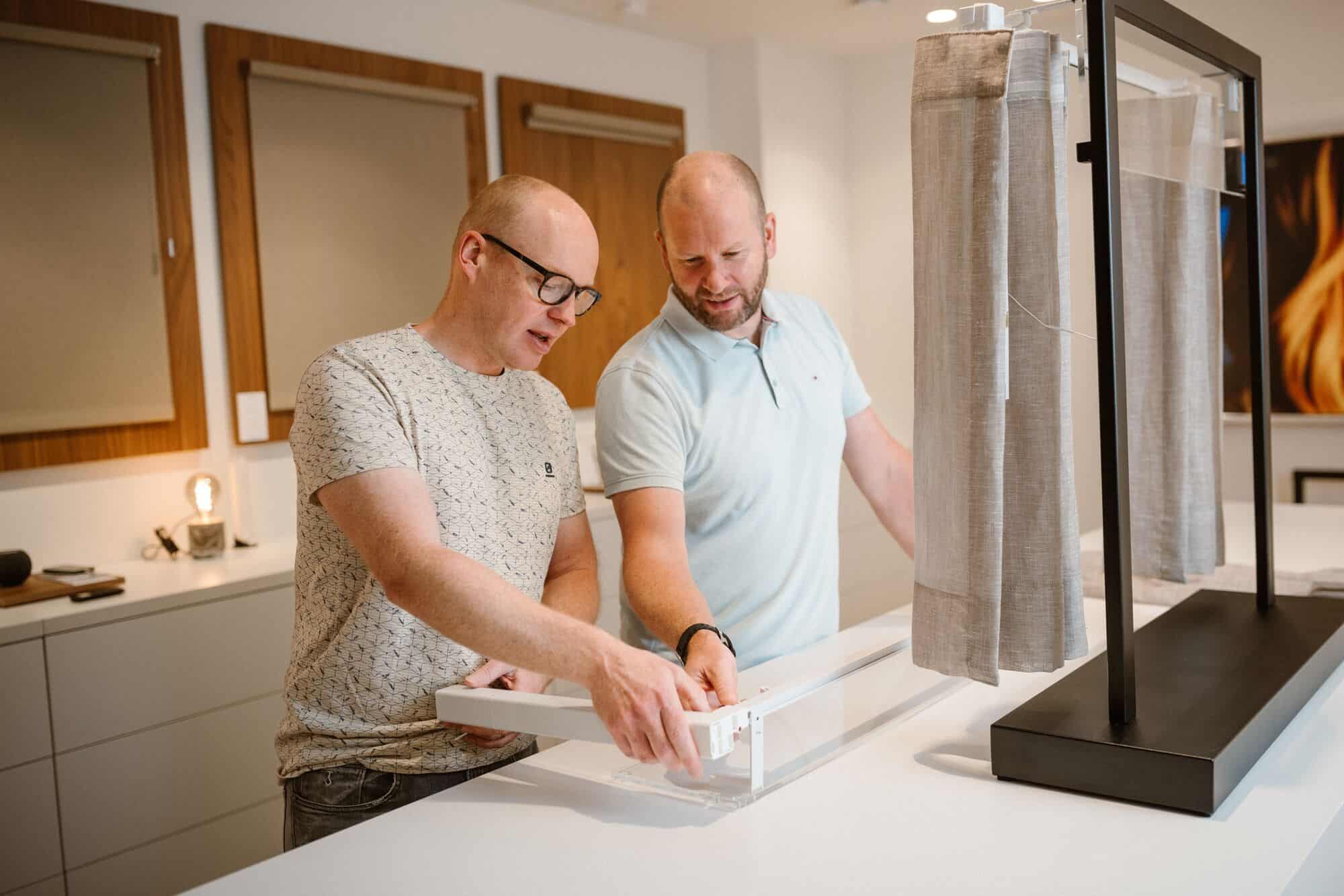 Two men stand at a counter assembling or examining a window blind or curtain rail in a modern, well-lit room with framed fabric samples on the wall behind them.