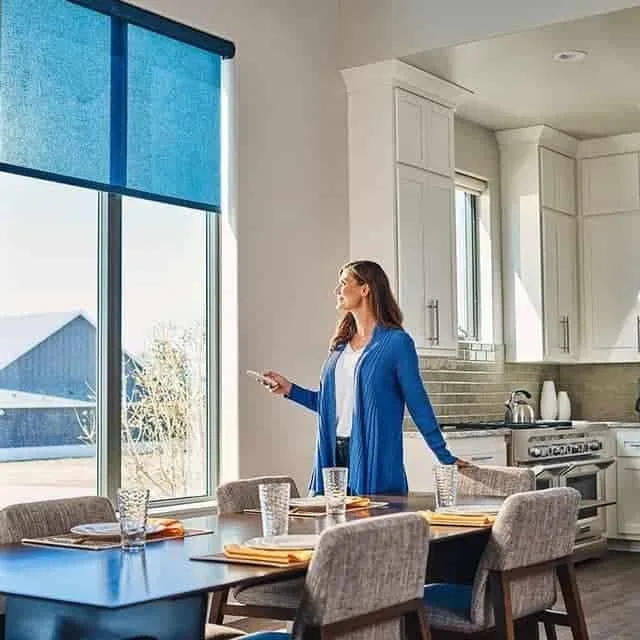 Woman standing in a modern kitchen using a remote to adjust window blinds.