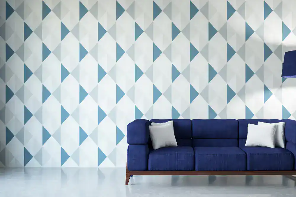 A modern living room in Broward County with a blue sofa and two white pillows, set against a wallpapered wall featuring a geometric pattern of blue and white triangles—showcasing thoughtful interior exterior custom design. A blue floor lamp stands on the right.