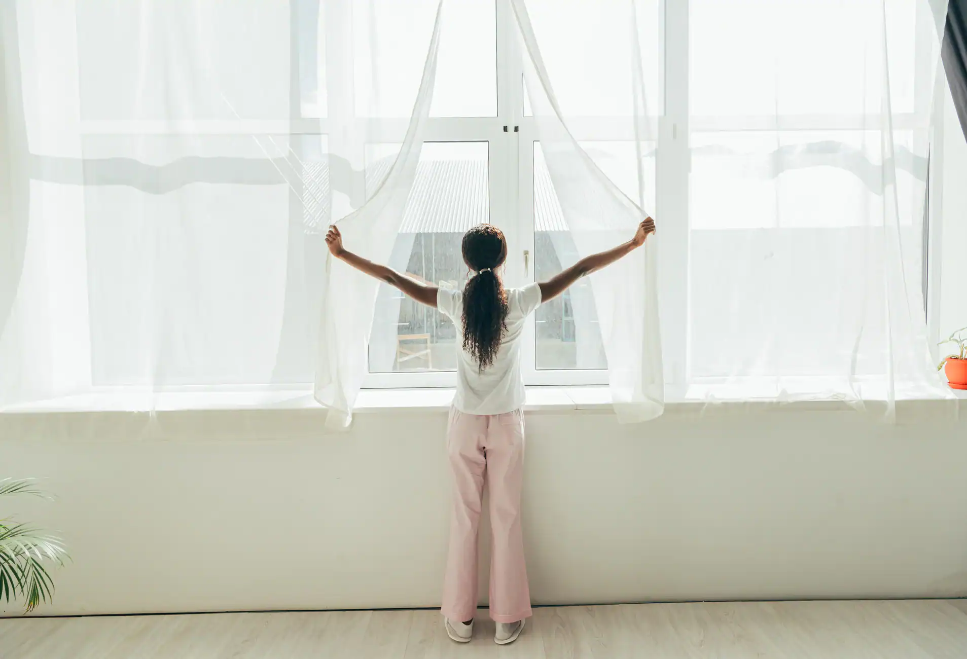 A person with long hair, wearing a white top and pink pants, stands facing a large window in a bright, minimalistic room in Broward County, holding sheer curtains open to let in sunlight. A plant adds charm to the custom design interior.