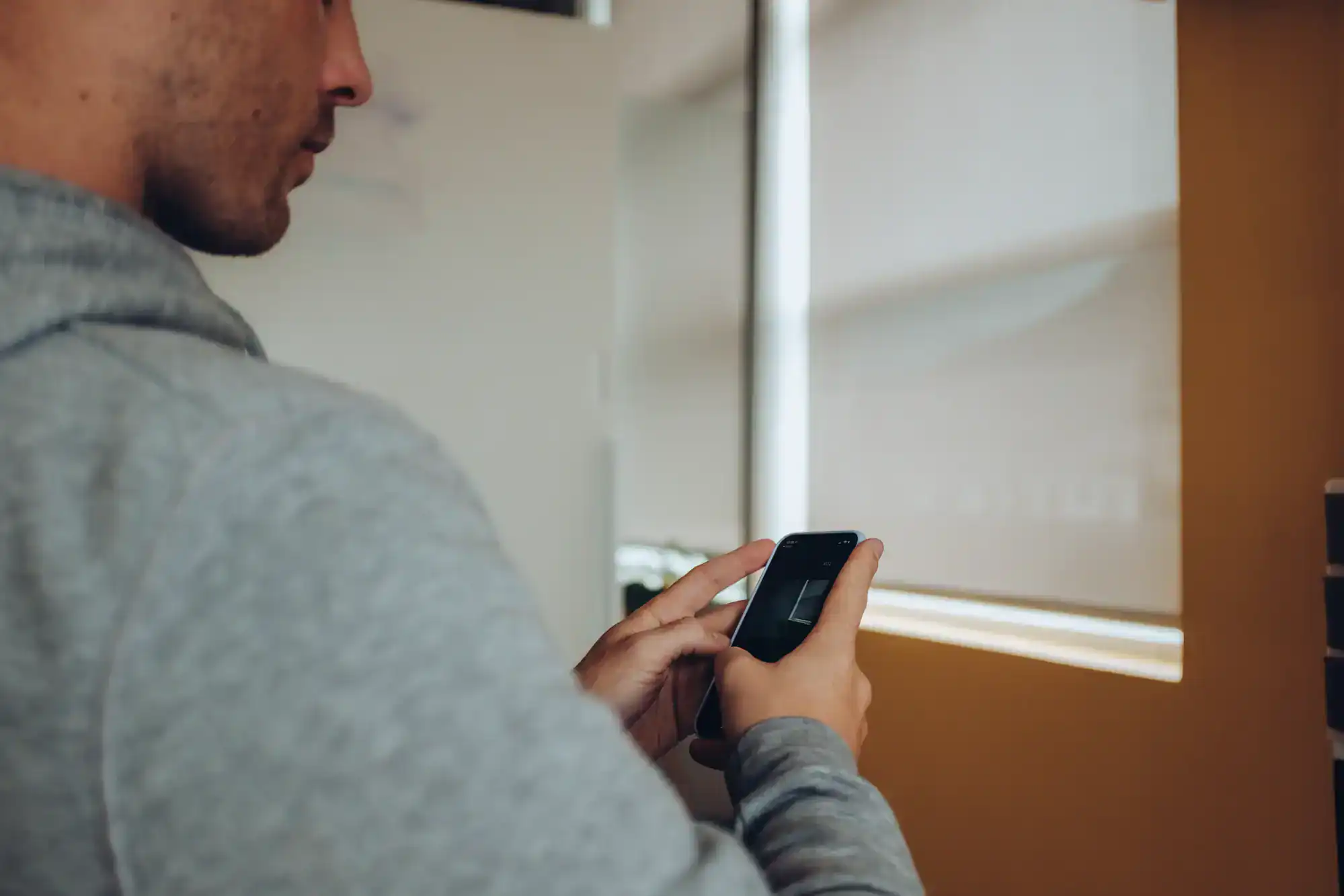 A person in a gray hoodie is holding and using a smartphone, standing indoors near large windows with blinds—highlighting Interior Exterior Custom Design details. The focus is on the hands and device.