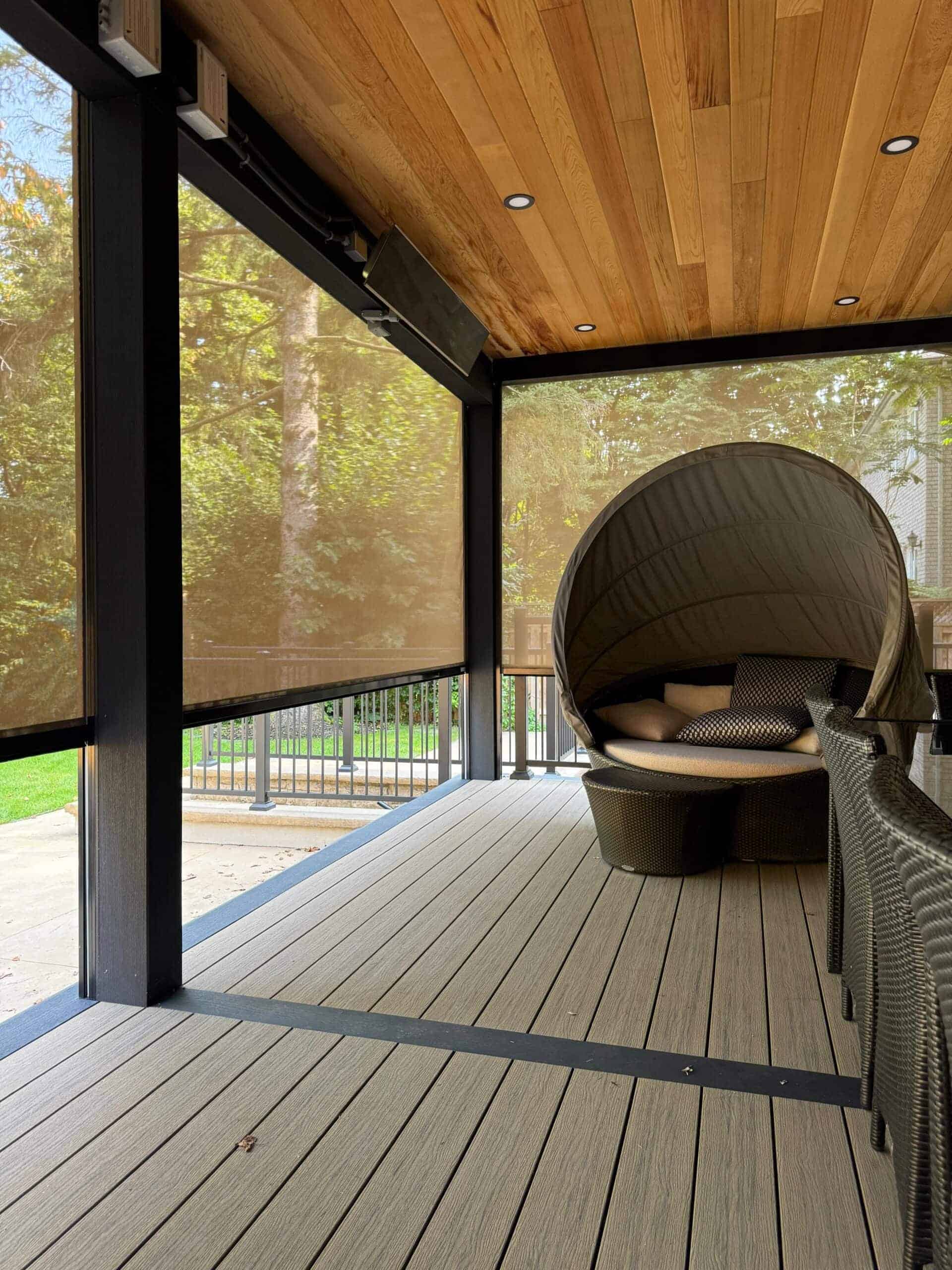 A covered outdoor patio with wood ceiling, retractable mesh shades, and composite deck flooring showcases Interior Exterior Custom Design. A round wicker lounge chair with cushions and canopy sits surrounded by trees in scenic Broward County.