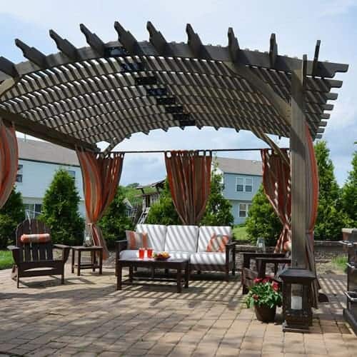Outdoor patio with a curved wooden pergola, striped orange curtains, cushioned seating, and a coffee table with drinks—an inviting example of Interior Exterior Custom Design in Broward County, surrounded by houses, trees, and potted plants.