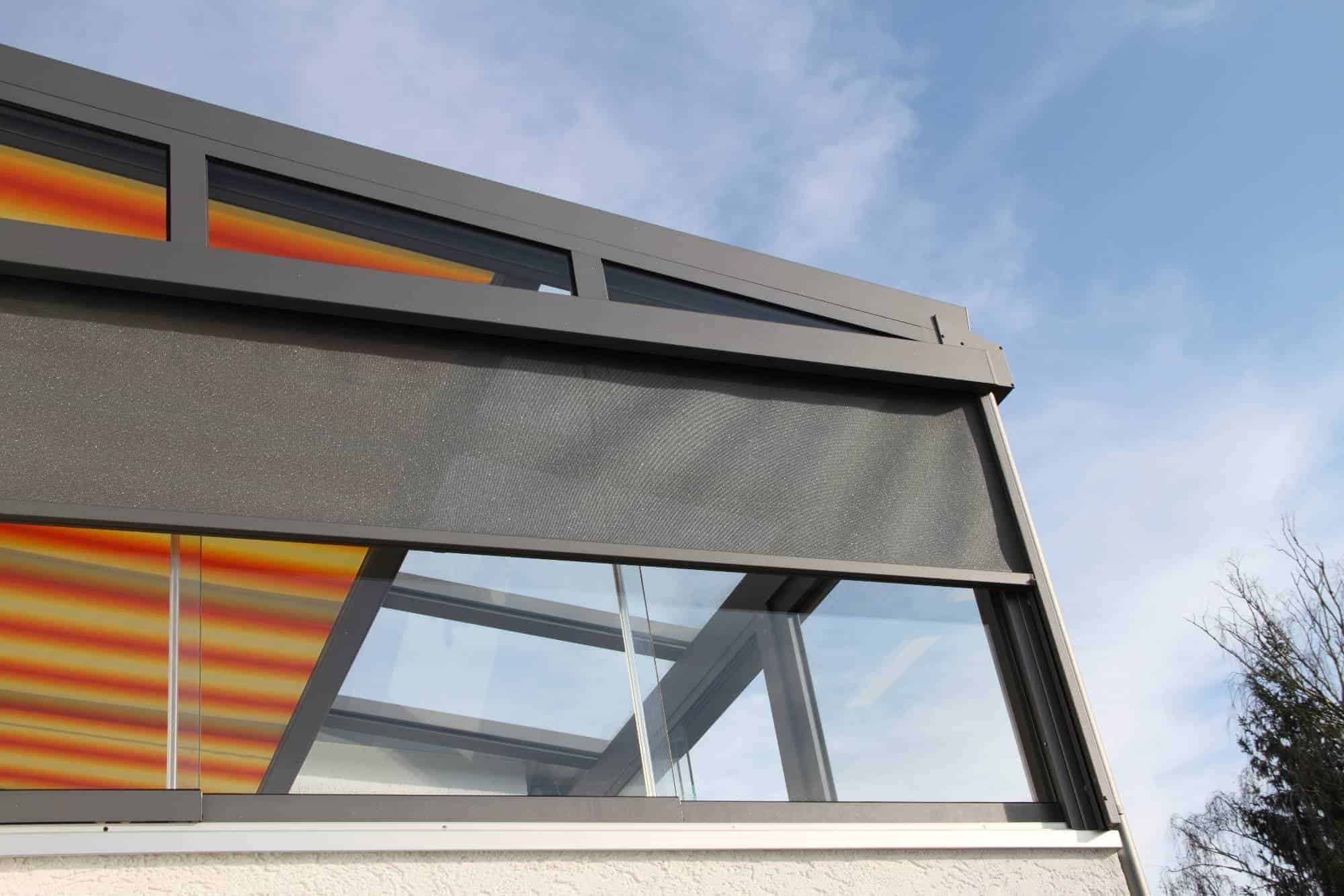 A close-up of a modern building facade in Broward County with large glass windows, a gray frame, and a partially lowered exterior roller shade, reflecting Interior Exterior Custom Design against a blue sky with light clouds.