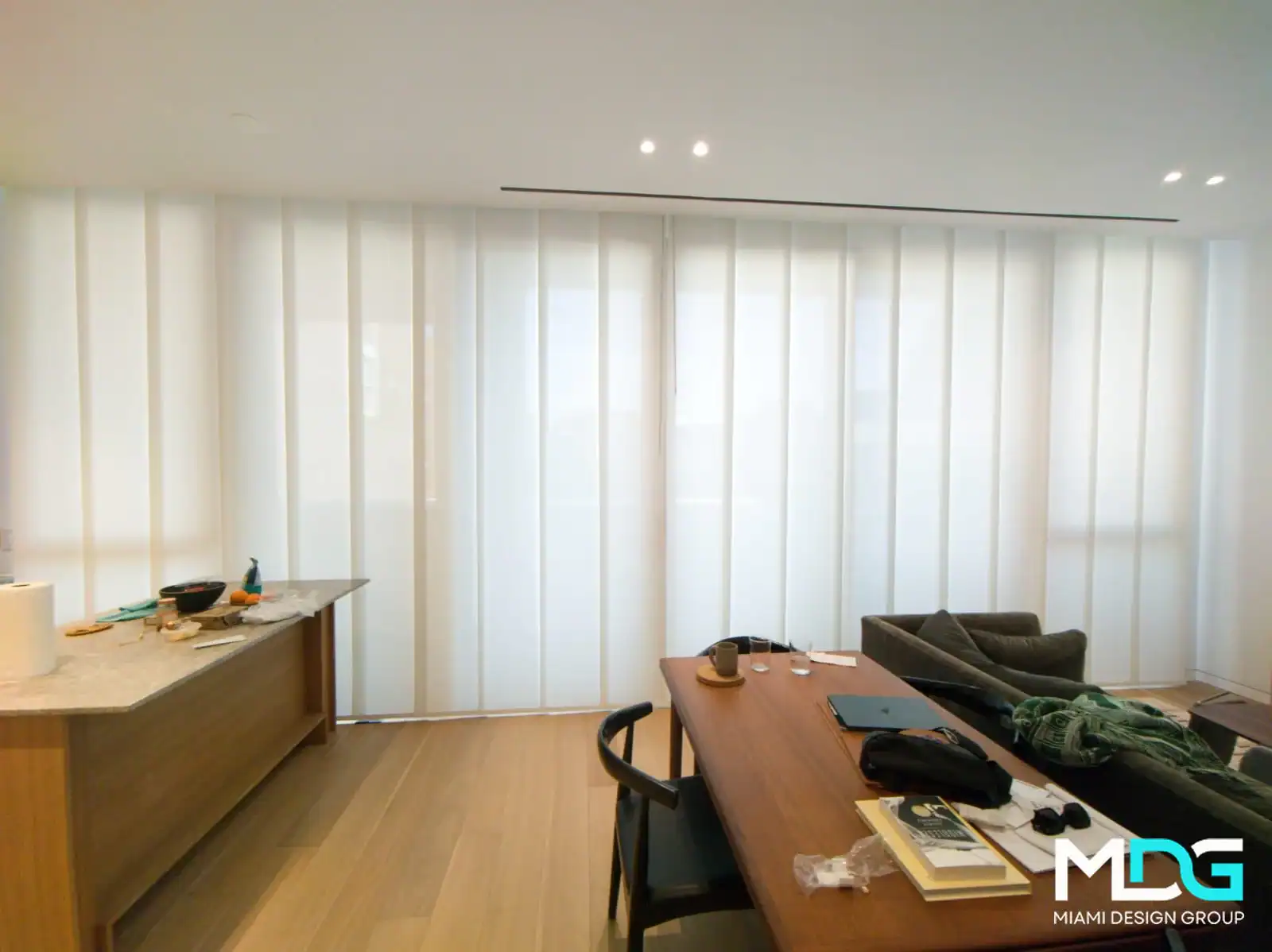 A modern living space in Broward County features floor-to-ceiling sheer white curtains, a wooden dining table with various items, a dark chair, and a kitchen counter with dishes. The MDG Miami Design Group logo appears in the bottom right corner.