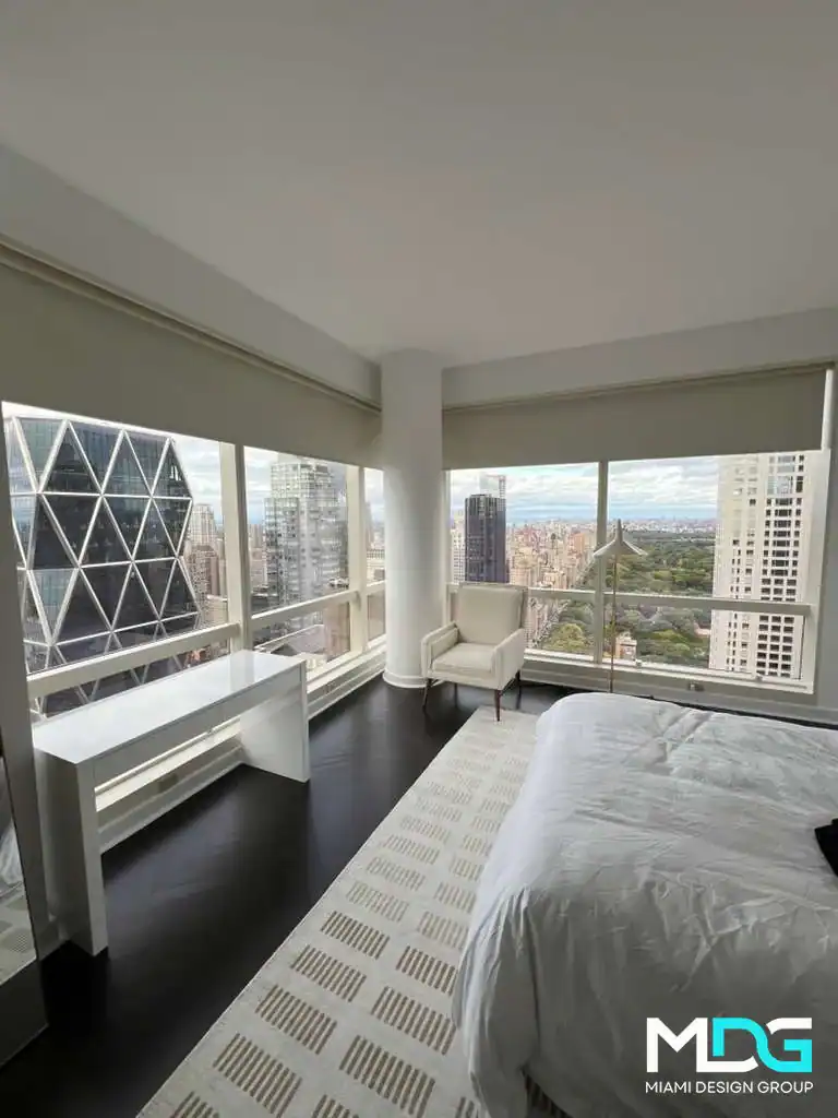 A modern bedroom with large floor-to-ceiling windows offering a cityscape view in Broward County. The room features a white bed, sleek desk, and dark wood floors&mdash;an example of Miami Design Group's Interior Exterior Custom Design.