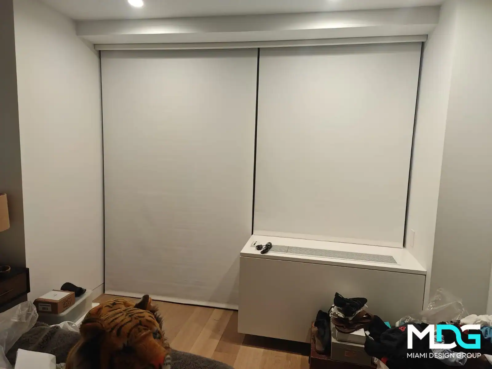 A modern, minimal bedroom by Miami Design Group in Broward County features white roller blinds on two windows, a white storage bench, plush tiger toy on the bed, and shoes on the wooden floor. Interior Exterior Custom Design touches complete the look.