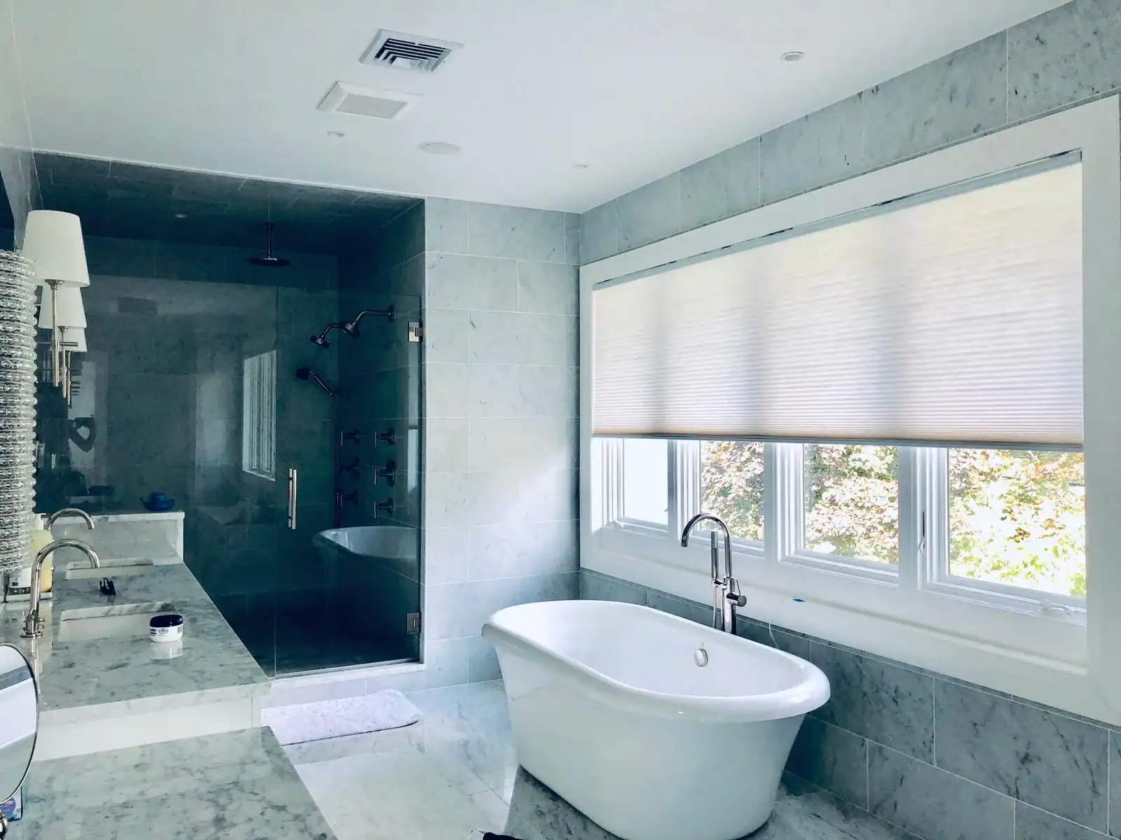 Modern bathroom with a freestanding white bathtub beneath large windows with blinds, marble countertops and walls, double sinks, and a glass-enclosed shower&mdash;showcasing Interior Exterior Custom Design excellence in Broward County.