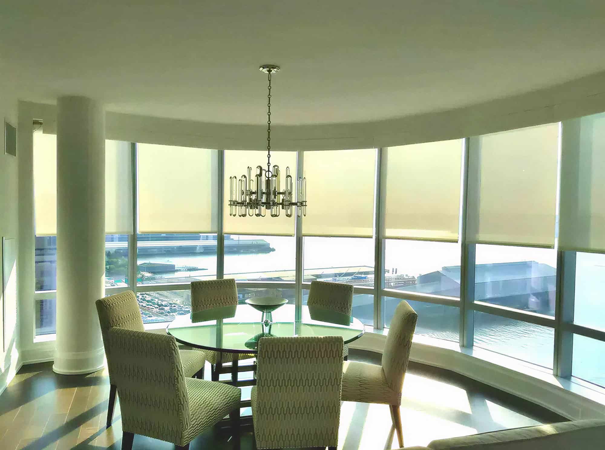 A modern dining area by large curved windows showcases a round glass table and six patterned chairs, with bright views of water and the Broward County cityscape, all enhanced by thoughtful Interior Exterior Custom Design touches.