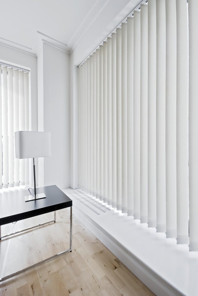 A modern, minimalistic room with light wood floors, large windows covered by vertical white blinds, and a black desk with a white lamp. Bright natural light fills the space.