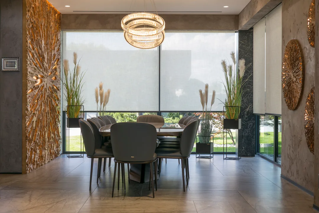 Dining area with pale shades across a broad window, a round table and sculptural pendant