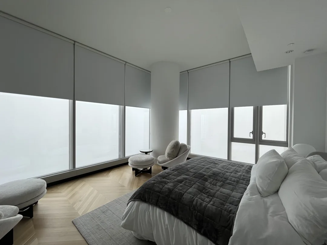 Corner bedroom with pale roller shades, lounge chairs and a dark quilted bed